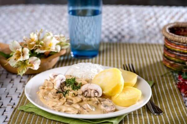 Chicken a la king on a white plate on top of a brown placemat with glass of water and flowers in the background